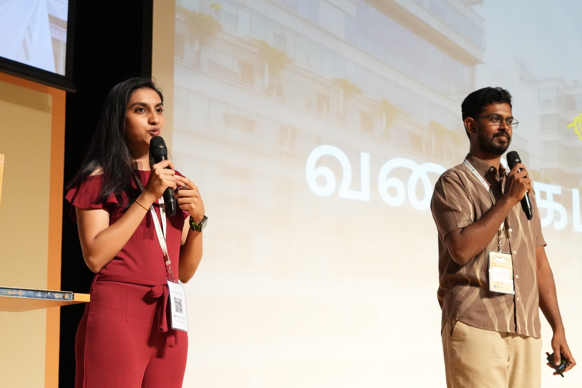 Two hosts welcoming the room in front of a Tamil greeting slide.